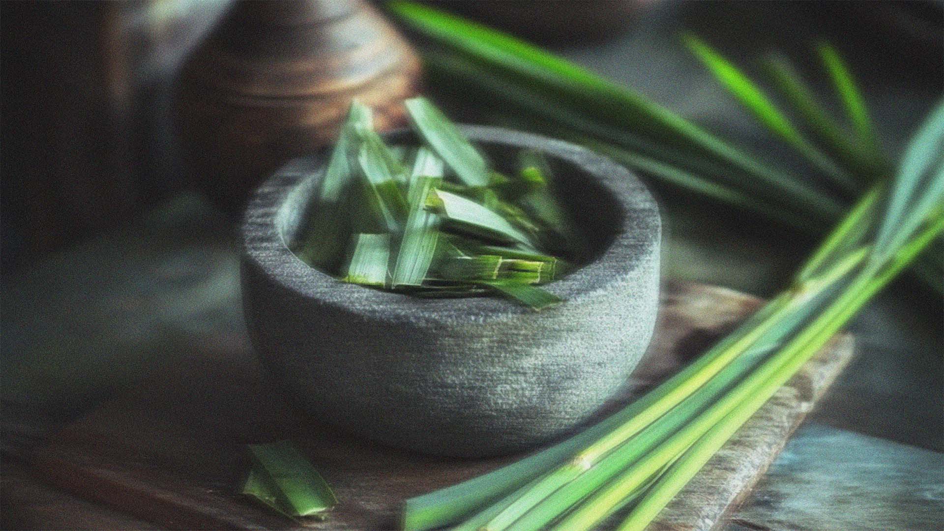 A stone made bowl full of pandan leaves with lemongrass stems on the side on a traditional kitchen setting.