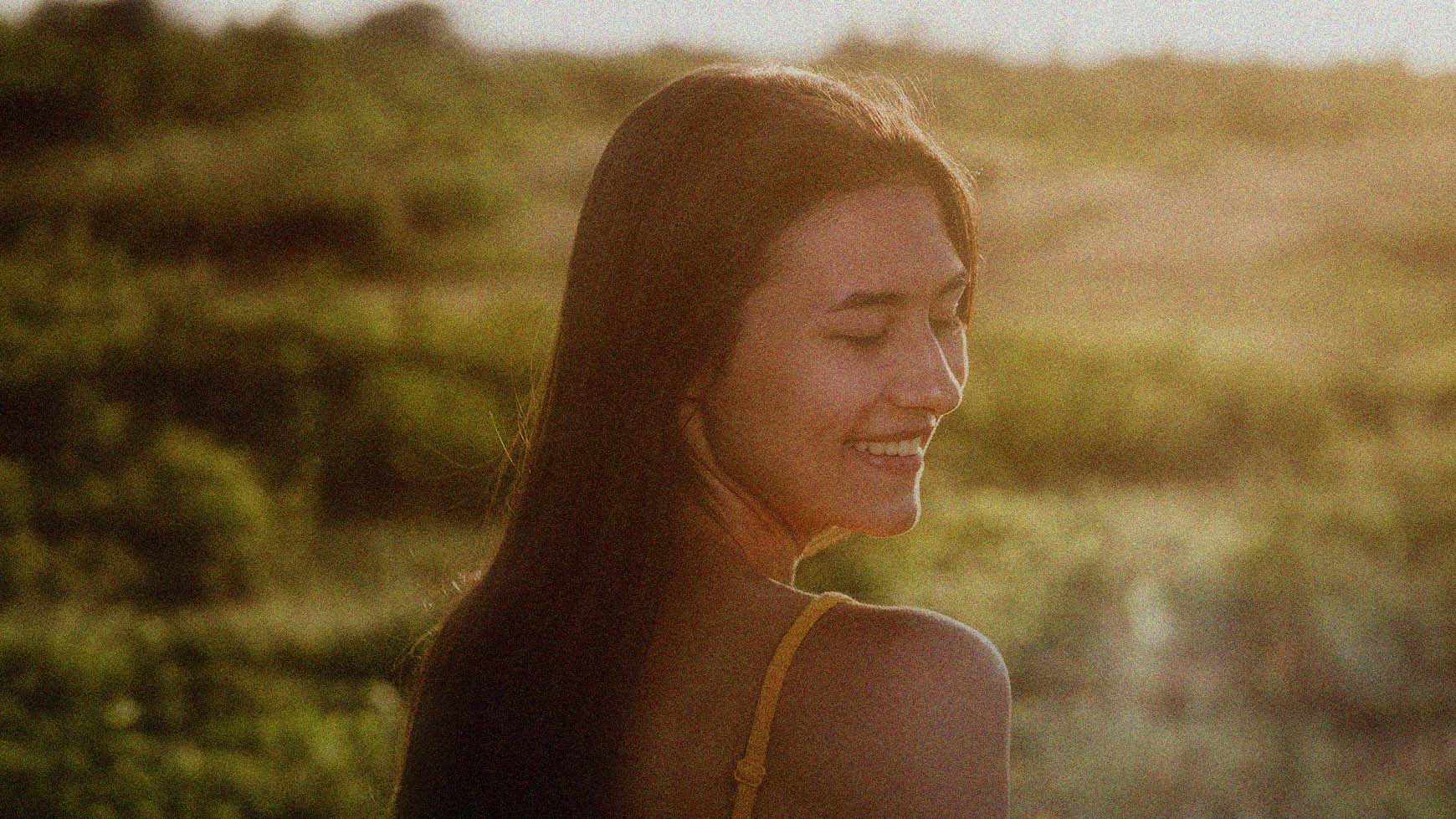 Woman with long hair smiling while standing in a field with a warm, golden light.