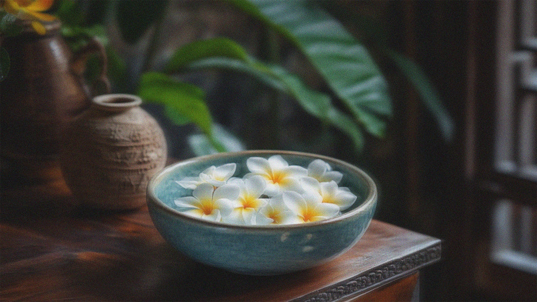 A porcelain bowl full of white frangipani flowers on a wooden table in a traditional Southeast Asian kitchen setting.