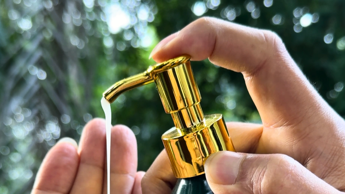 A bottle of hand and body wash being held between two hands, pumping wash out of a golden pump over a natural background.