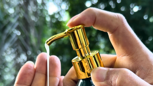 A bottle of hand and body wash being held between two hands, pumping wash out of a golden pump over a natural background.