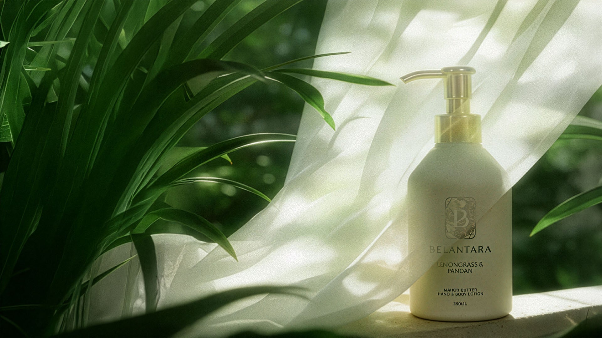 A bottle of the Belantara Mango Butter Hand & Body Lotion partially covered by a white veil surrounded by pandan leaves. 