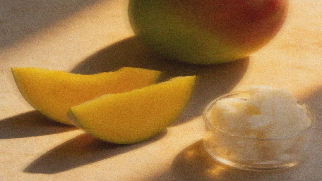 A mango fruit, 2 slices of mango and a glass container with mango butter inside, under indirect sunlight.