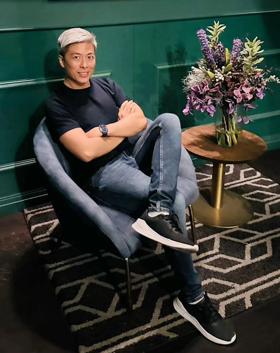 Shawn, the founder of Belantara, wearing black t-shirt and jeans sitting in an opulent lounge, featuring a chair and flowers on a side table.